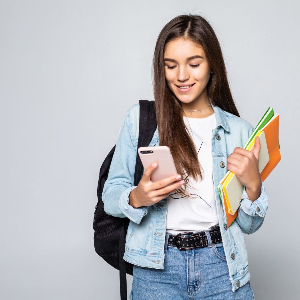 portrait-happy-young-woman-standing-with-backpack-holding-books-mobile-phone-isolated-white-wall (1)